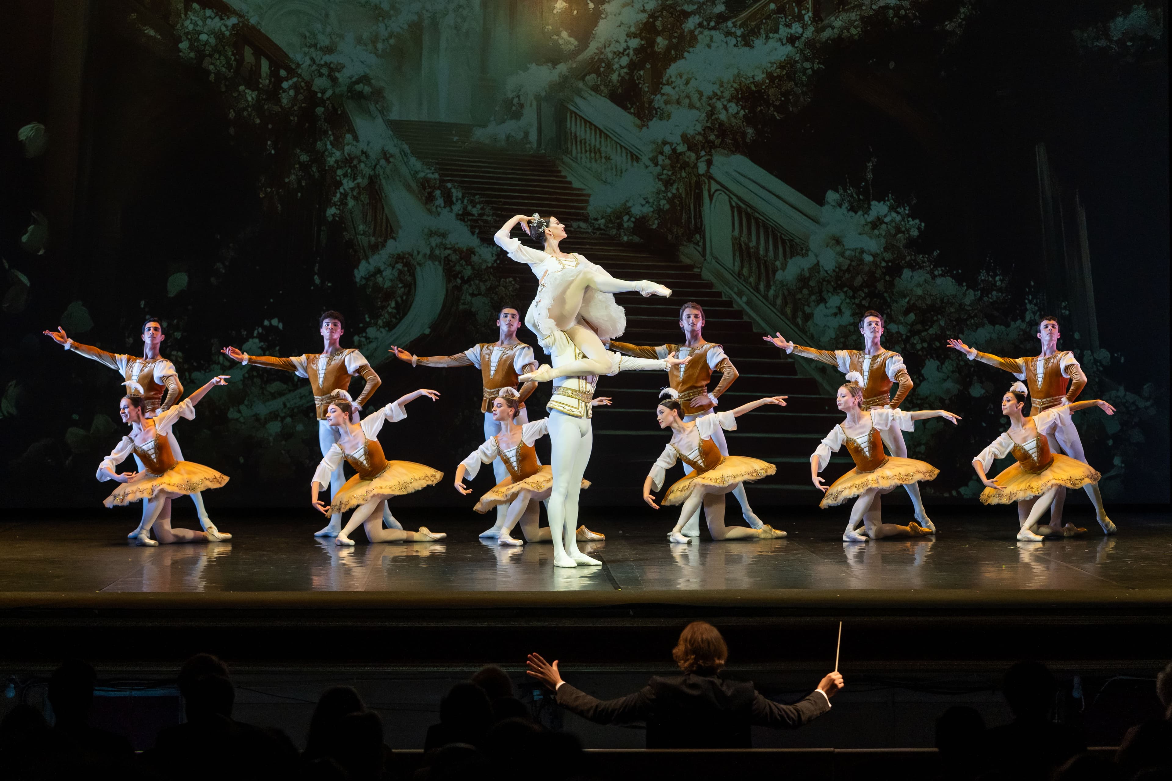 Ballet corps in gold and white costumes on stage with a dancer lifted at the centre, conductor's hands below
