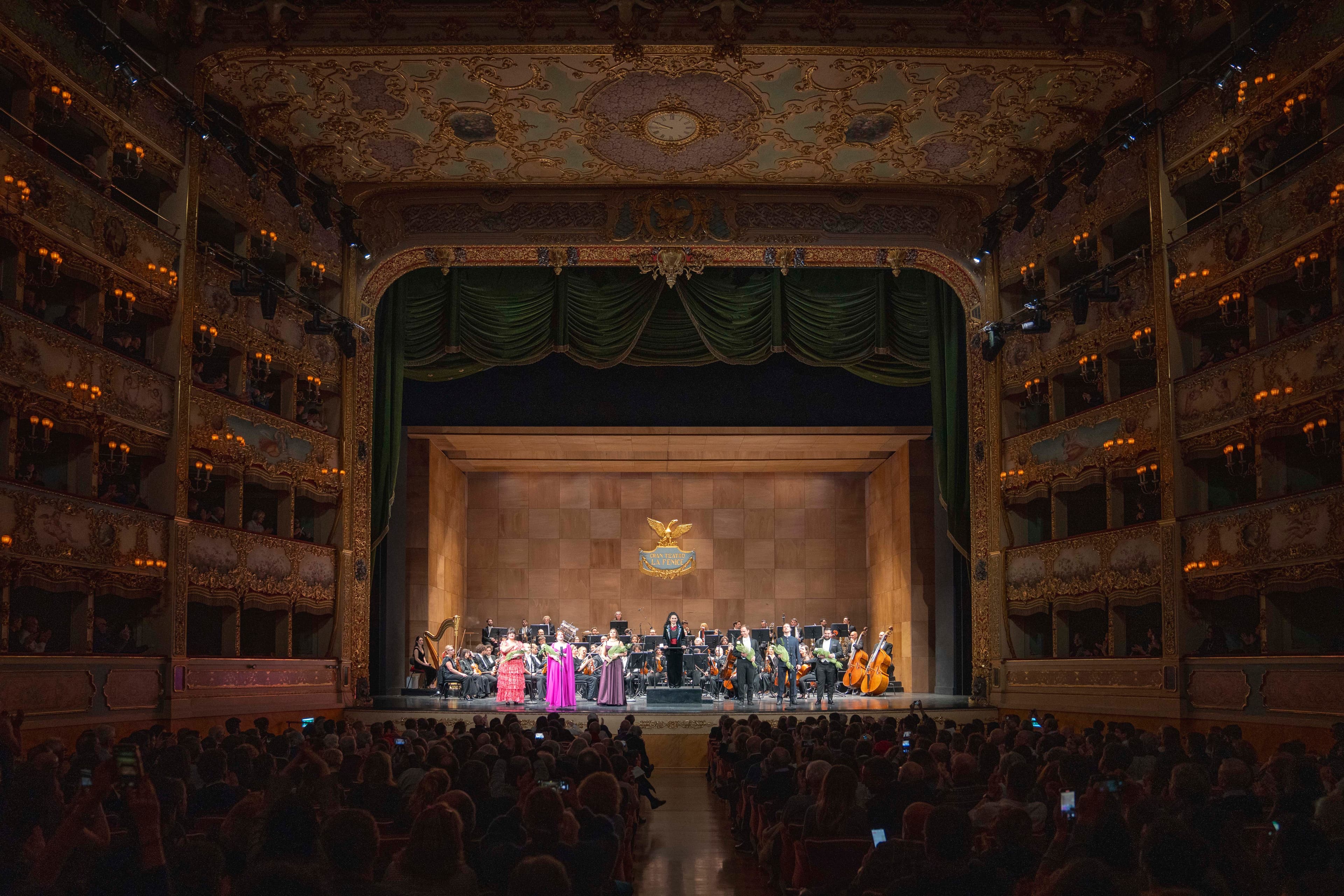 Orchestra and soloists on the stage of a historic horseshoe-shaped opera house, audience in the foreground
